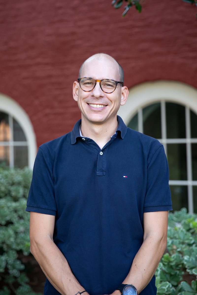 Photo of Nicolas Delon wearing glasses, a blue polo shirt, red bricks and a leaves in the background